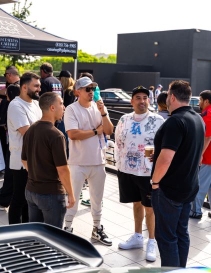 A gathering of stylishly dressed men in hats and casual wear conversing outdoors under a black canopy.
