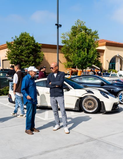 Two men stand talking in a parking lot, with a white sports car and other vehicles behind them.