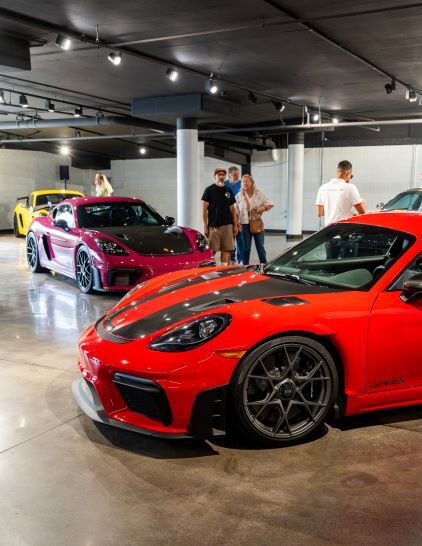 A red Porsche Cayman sits in a showroom, while a few people observe classic cars nearby.
