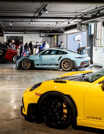 A light blue Porsche 911 GT3 RS sits in a showroom surrounded by other cars and people looking at them.