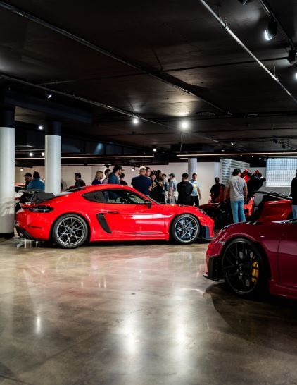 A shiny red Porsche sits on display in a dimly lit showroom surrounded by onlookers.