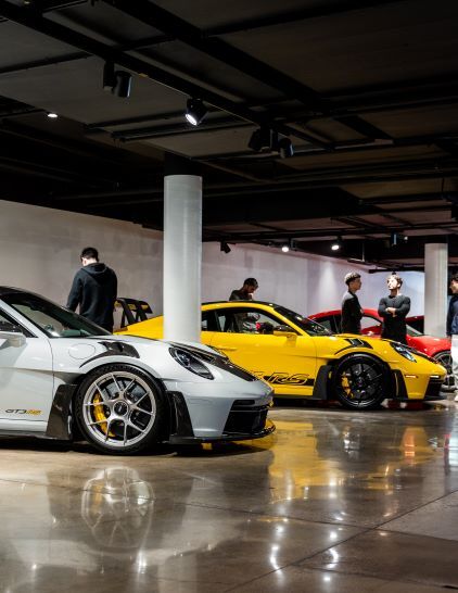 Several Porsche cars in various colors are displayed in a showroom with people observing them.