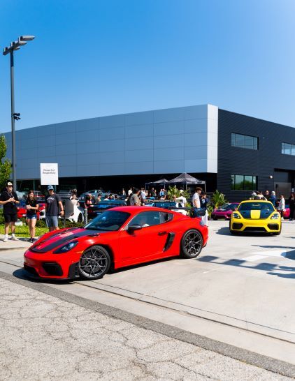 A bright red sports car sits parked at a car show with a yellow car nearby and people admiring the vehicles.