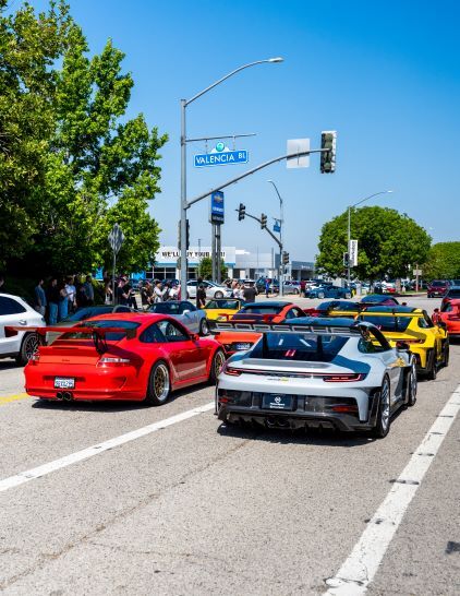 A line of luxury sports cars in various colors parades down a sunny city street.
