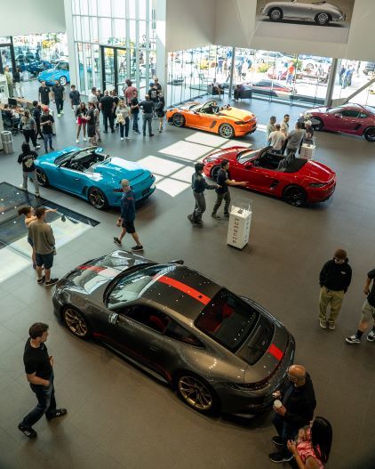 Several people browse luxury cars displayed in a modern showroom with large windows overlooking an outdoor lot.