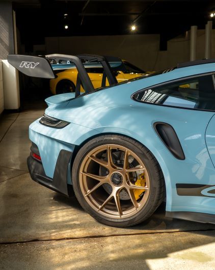 A blue Porsche 911 GT3 RS sits in a dimly lit garage with a yellow sports car in the background.