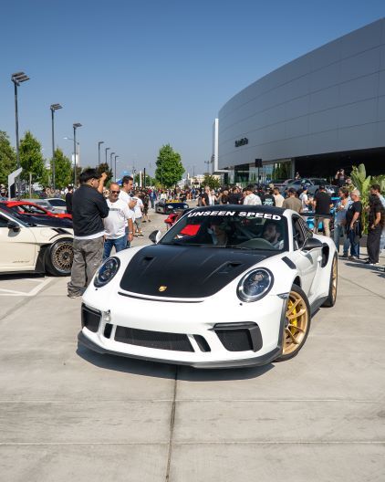 A black and white Porsche 911 sits in a crowd at a car show, with onlookers taking photos.