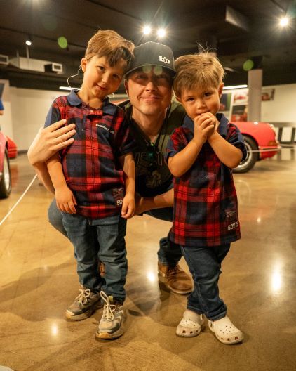 A man kneels, smiling with his arms around two young boys who pose for a picture.