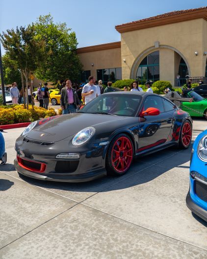 A grey Porsche 911 with red accents sits parked in front of a building during a car show.