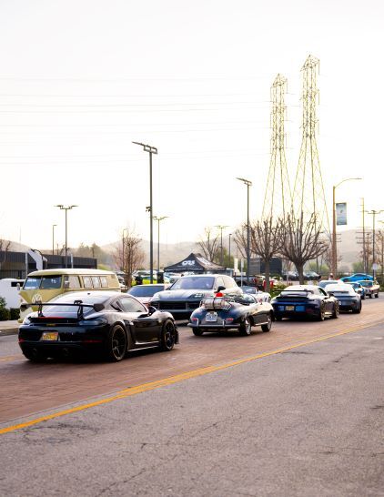 A line of dark-colored luxury cars drives along a street with transmission towers in the background.
