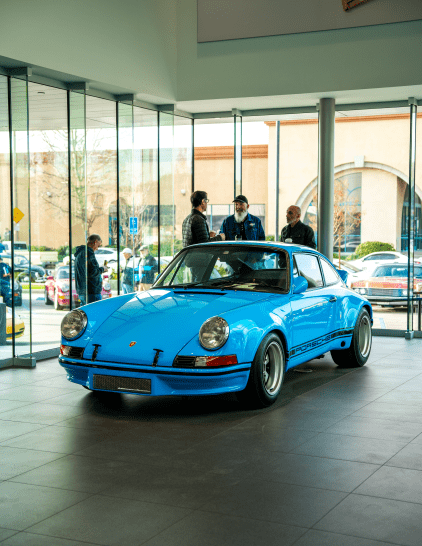 A bright blue classic Porsche sits inside a dealership showroom with people observing it.