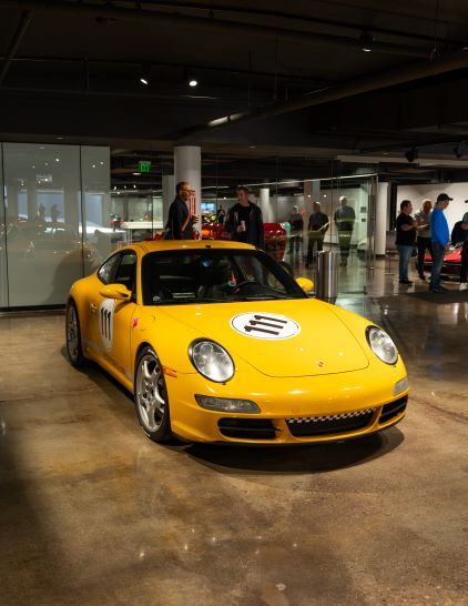 A yellow Porsche race car sits on display in a modern showroom with onlookers.