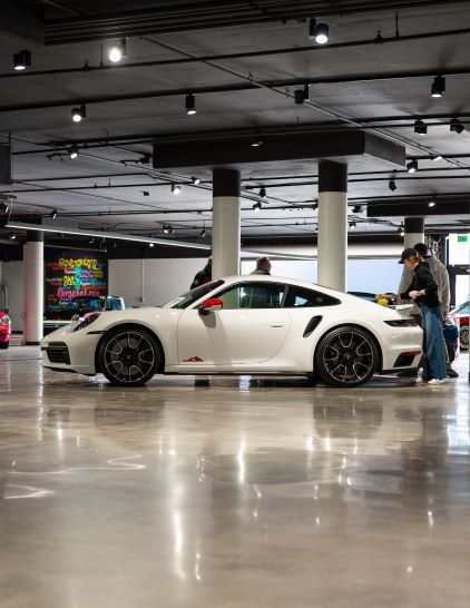 A white Porsche 911 sits on a polished floor as people observe it in a modern showroom.