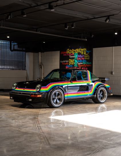 Black Porsche 911 with a rainbow stripe across the side, parked on a concrete floor.