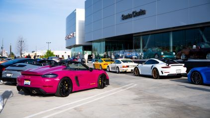 Several luxury cars, including a bright pink Porsche, are displayed in a car dealership parking lot.