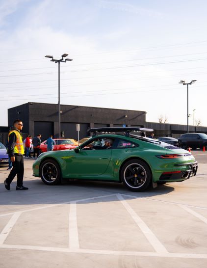 A green Porsche 911 drives in a parking lot with a man in a yellow vest nearby.