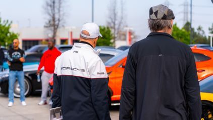 Two men in matching jackets and hats observe a line of luxury cars at an outdoor event.