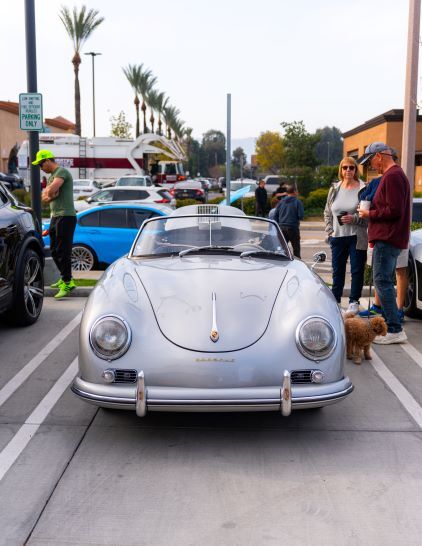 Front view of a silver vintage Porsche convertible surrounded by people and other cars in a parking lot.