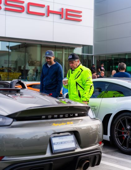 Two men in vests stand near a gray Porsche Spyder RS at a dealership.