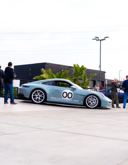 A light blue Porsche 911 with racing stripes is parked on pavement near onlookers and landscaping.