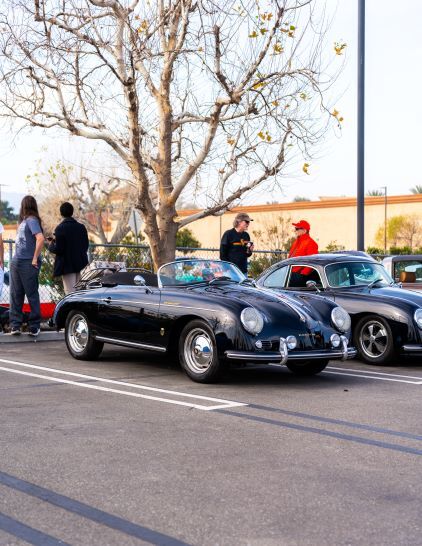 Several people examine shiny classic Porsche cars parked at an outdoor car show.