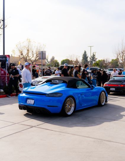 A bright blue Porsche 718 Boxster convertible sits at a car show surrounded by spectators.