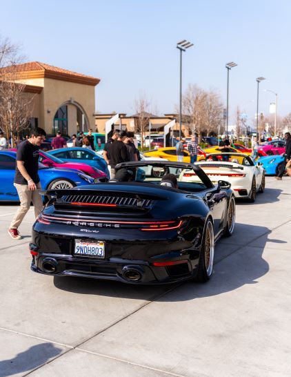 A black Porsche convertible displays itself at a car show with several other luxury vehicles nearby.