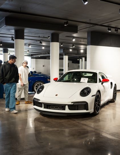 Two people examine a white Porsche 911 in a modern showroom with exposed ceiling beams.