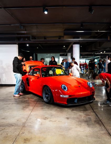A bright red Porsche 911 on display is admired by a crowd of people in an exhibition hall.