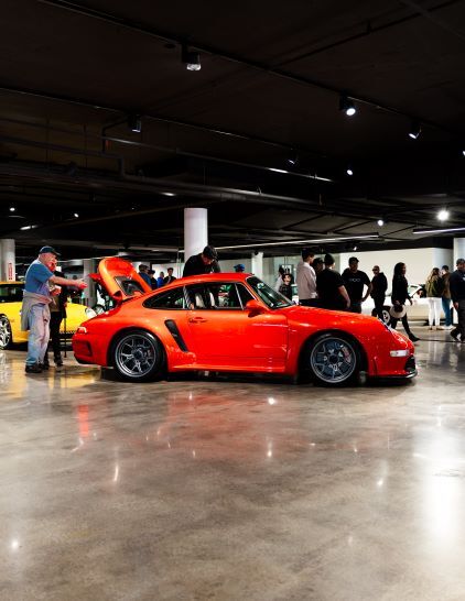 A man stands near a bright red Porsche with its hood open at a car show.