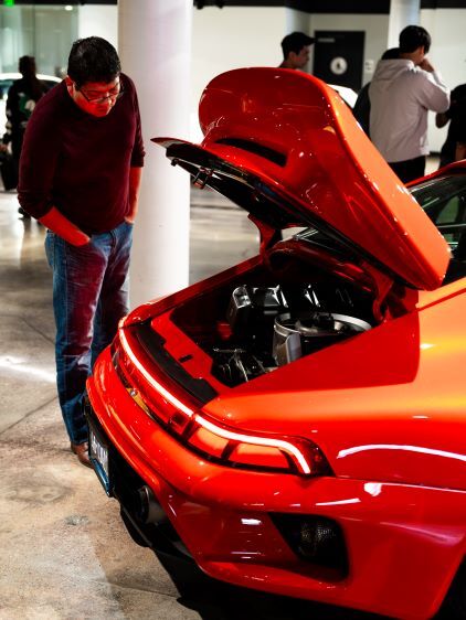 A man in glasses examines the engine of a vibrant orange sports car with its hood raised.