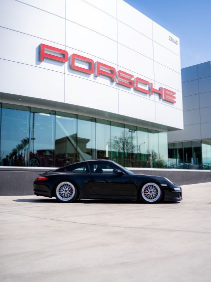 A black Porsche sports car sits parked in front of a modern Porsche dealership building.