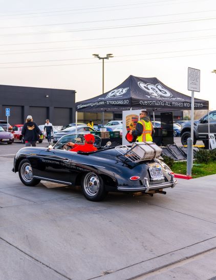 A person in a reflective vest stands near a classic silver Porsche convertible at an outdoor car show.