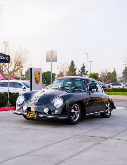 A black vintage Porsche Speedster sits parked on a street with a Porsche dealership sign visible in the background.