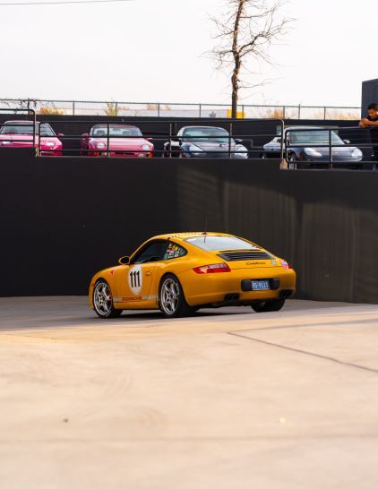 Yellow Porsche 911 race car with number 111 on the side, parked in a lot, with other cars above.