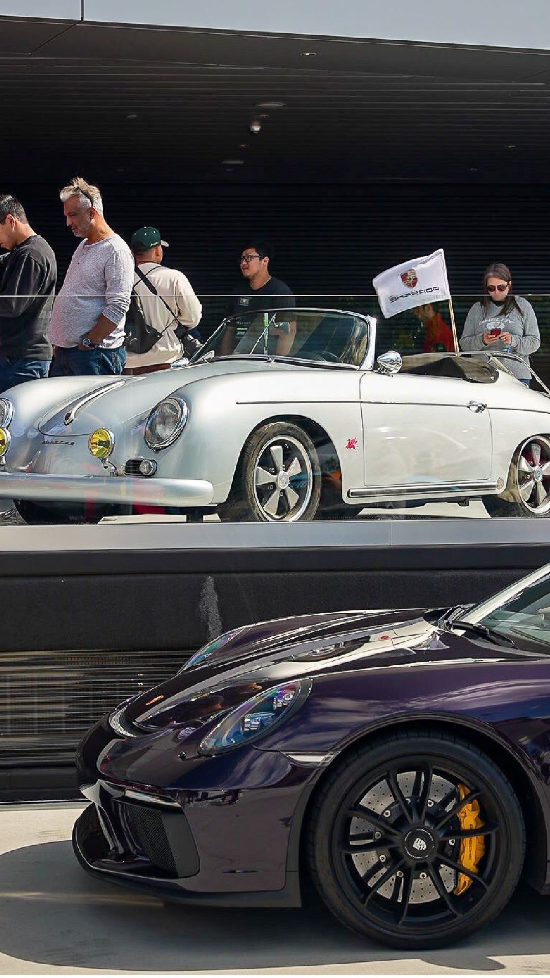 A silver vintage Porsche 356 Speedster convertible with a Porsche flag, and a dark modern Porsche sportscar.