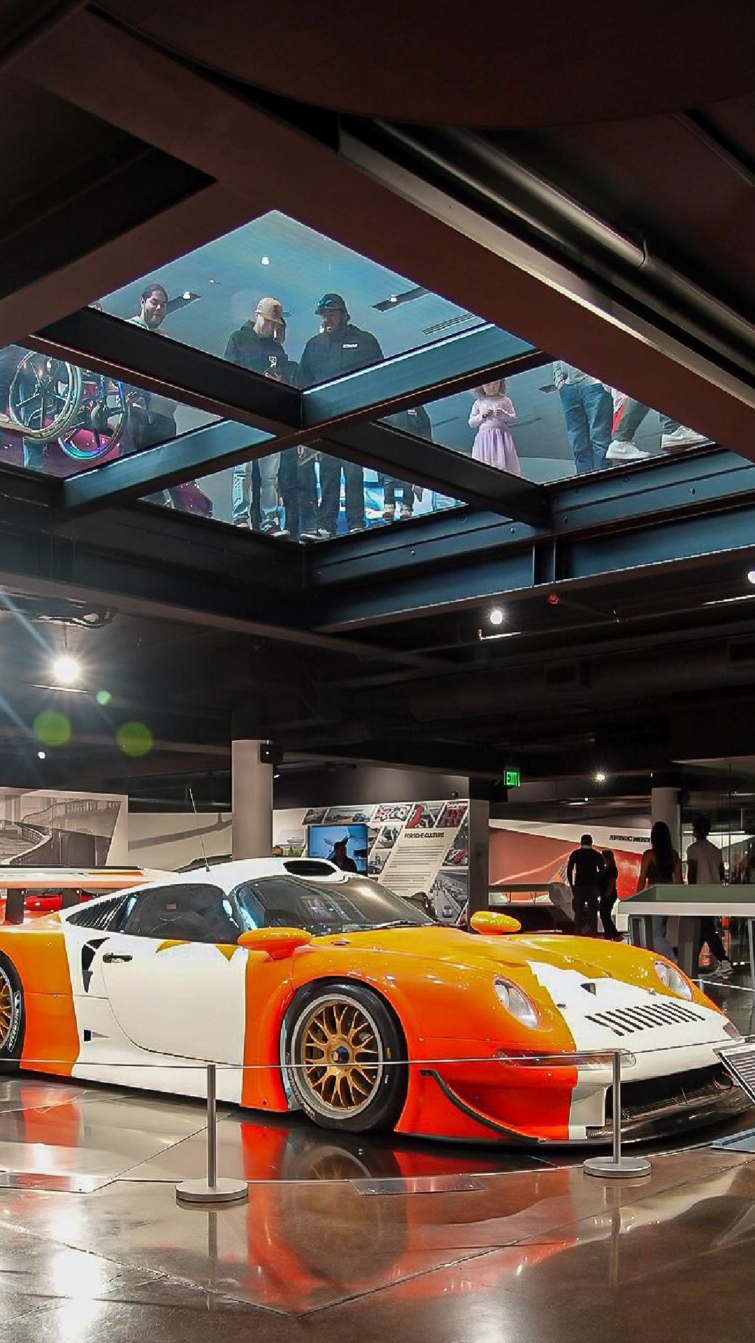 A futuristic orange and white race car on a polished floor, viewed from below a glass walkway with people.