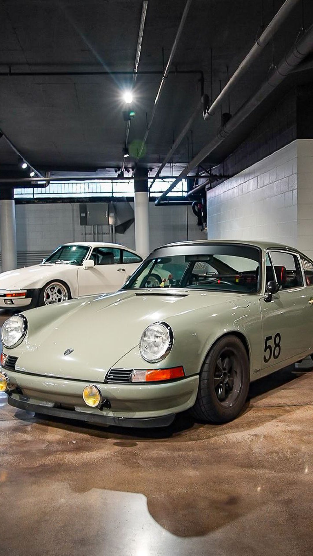 An olive green Porsche 911 with 58 on the side is parked next to a white Porsche 944 in a garage.