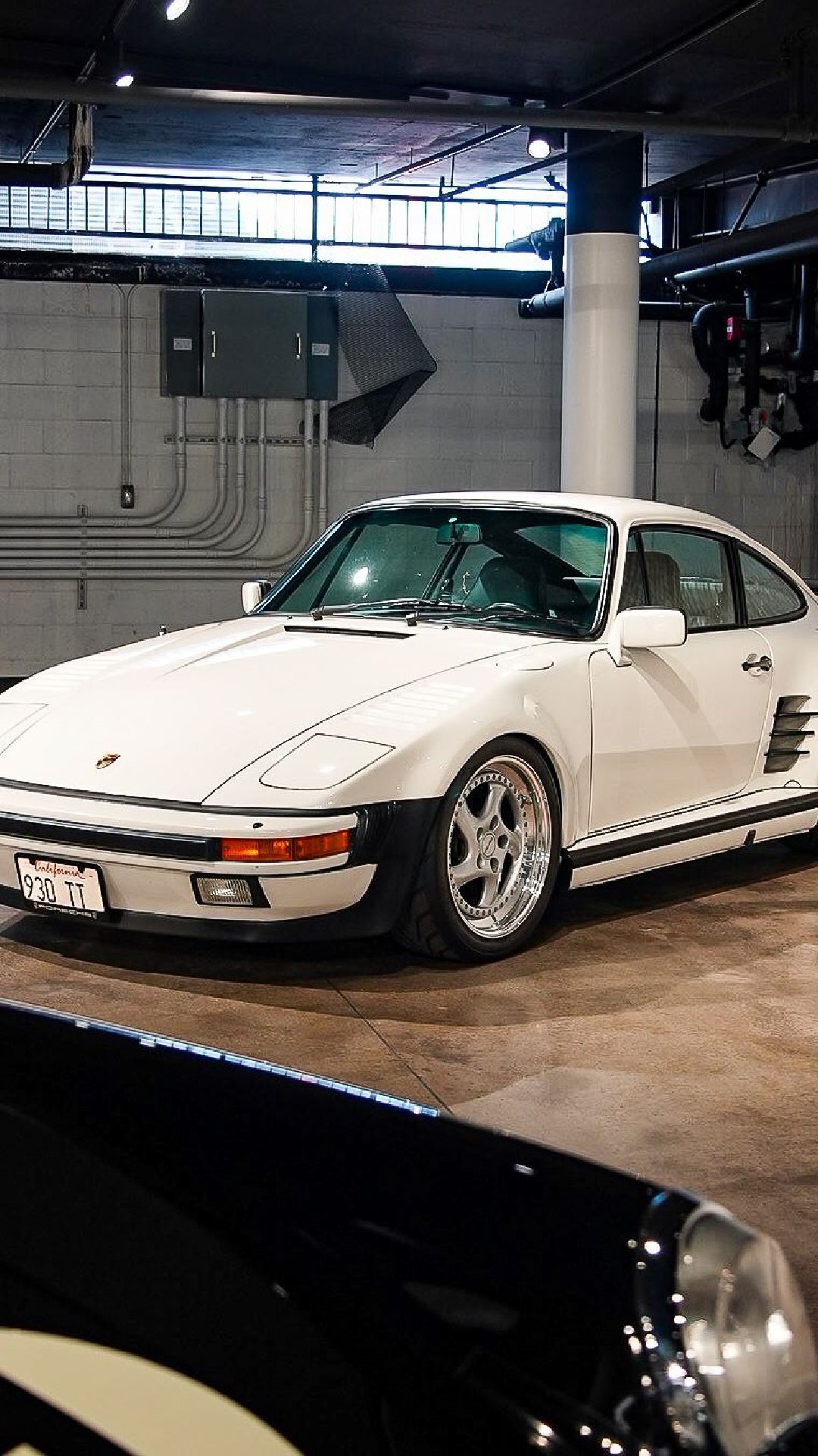 A white vintage Porsche 930 TT with shiny chrome wheels, parked in a dimly lit garage.