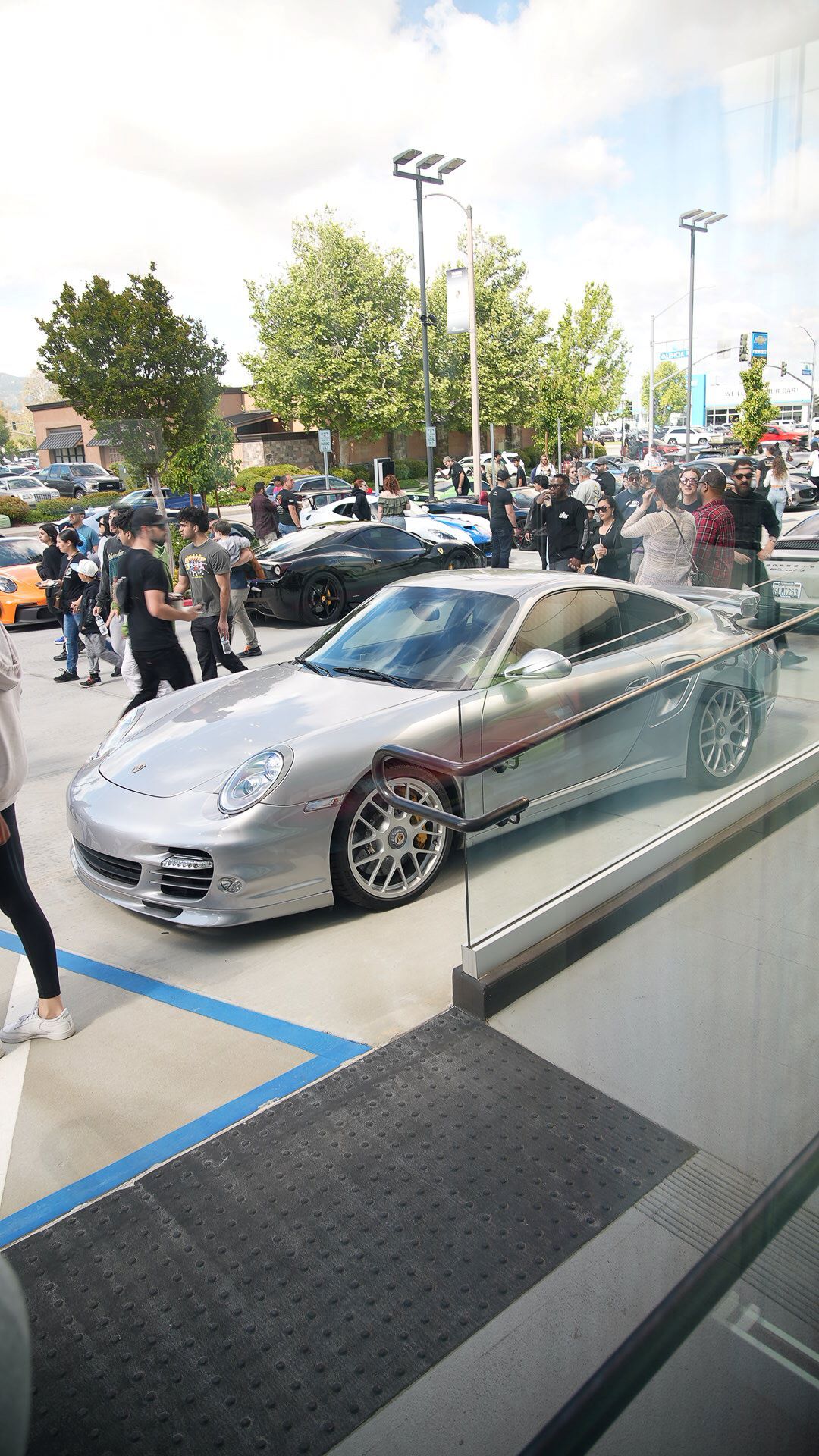 A silver Porsche, seen partially through a glass barrier, is parked on a paved lot with a crowd of people and other luxury cars.