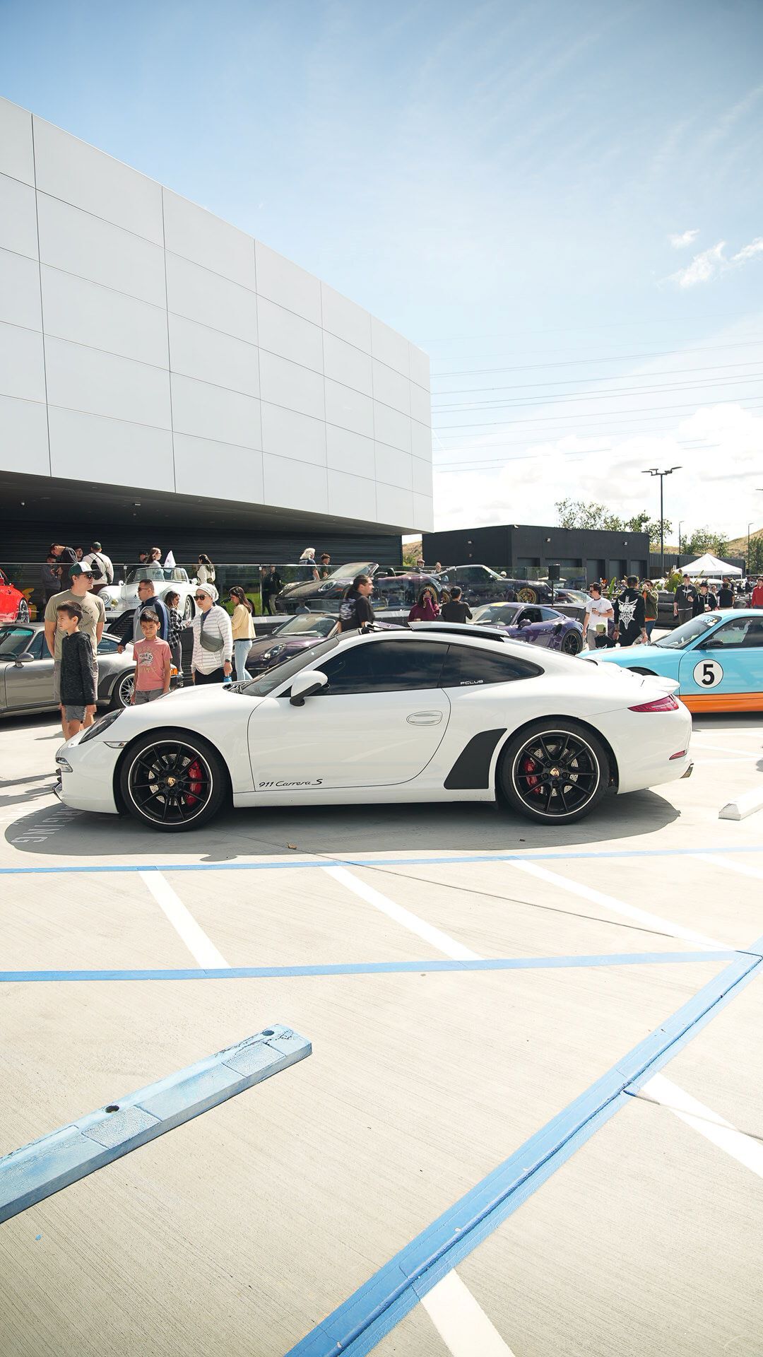 A white Porsche 911 Carrera S parked outdoors at a car show, with people and other cars in the background.