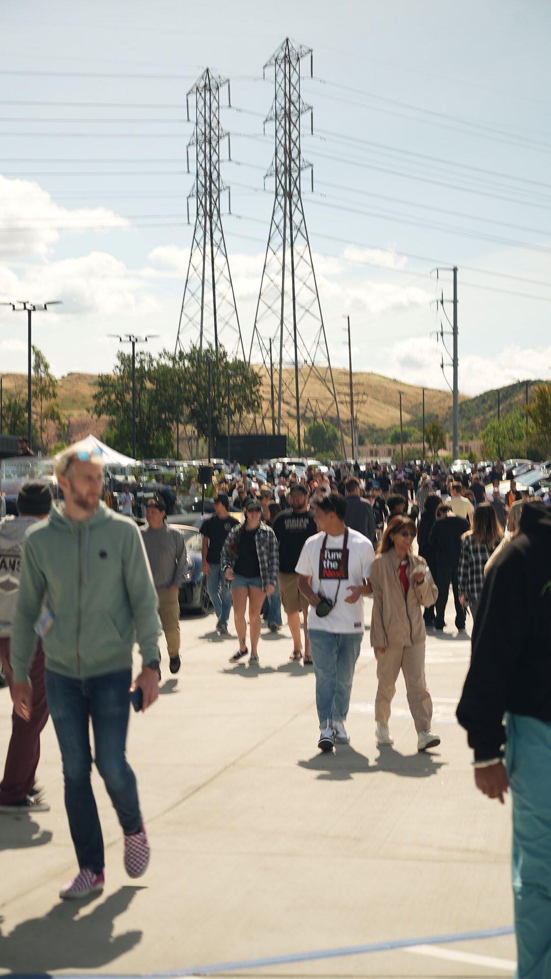 Attendees walk past power lines and hills at an outdoor event on a sunny day.