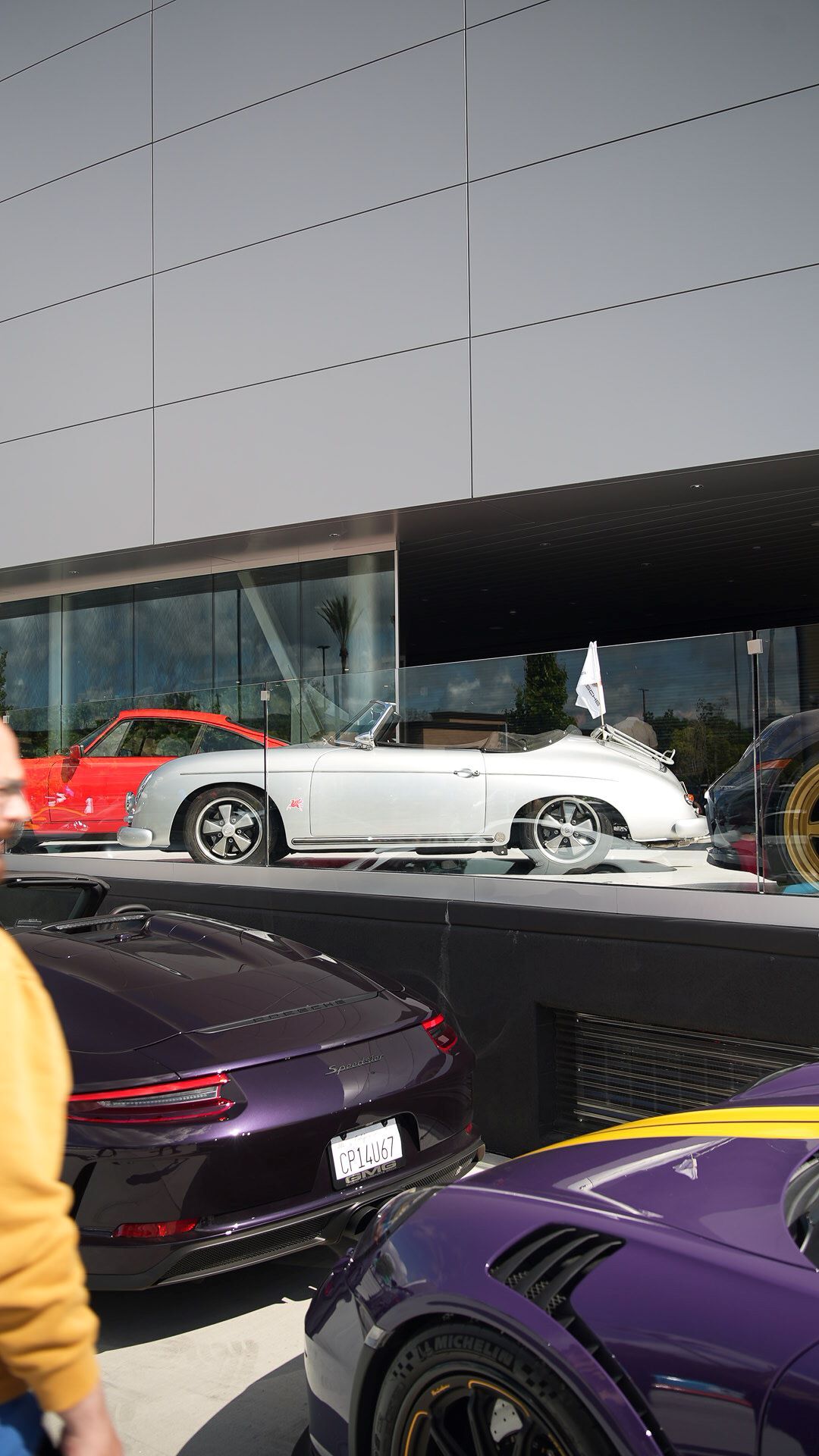 Three Porsche cars: a silver convertible behind glass, and two purple cars in the foreground.