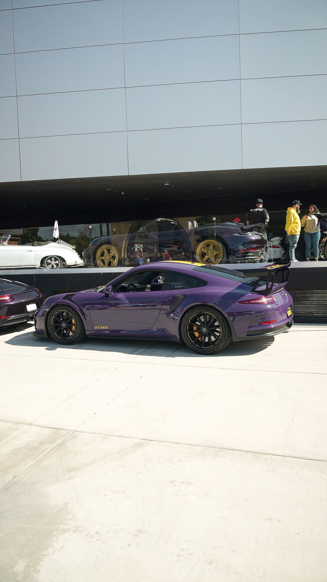 A purple Porsche GT3RS parked before a modern building with other Porsches displayed in a glass-walled showroom.