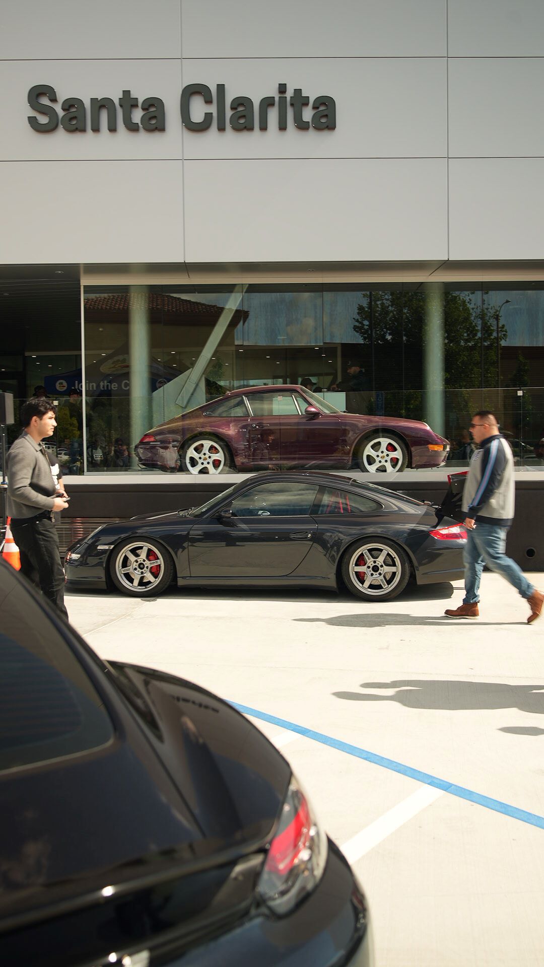 Two men near a black sports car parked outside a building with Santa Clarita on its facade. A purple sports car is visible inside the building's display window.