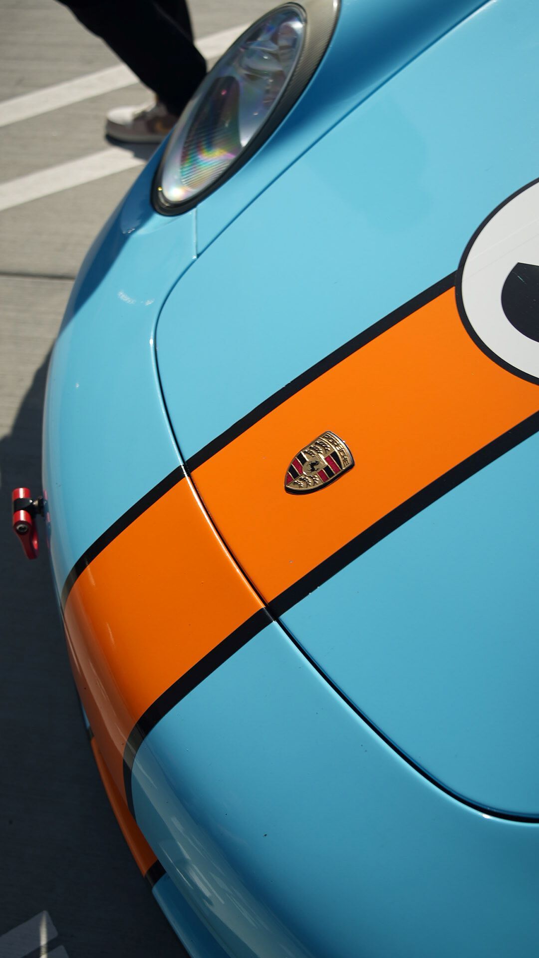 Close-up of a light blue racing Porsche with an orange racing stripe and black outline, with a detailed Porsche emblem.