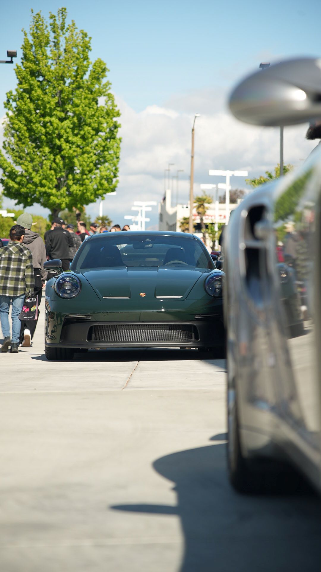 Front view of a dark green Porsche sports car parked outdoors with people and another car in the background.