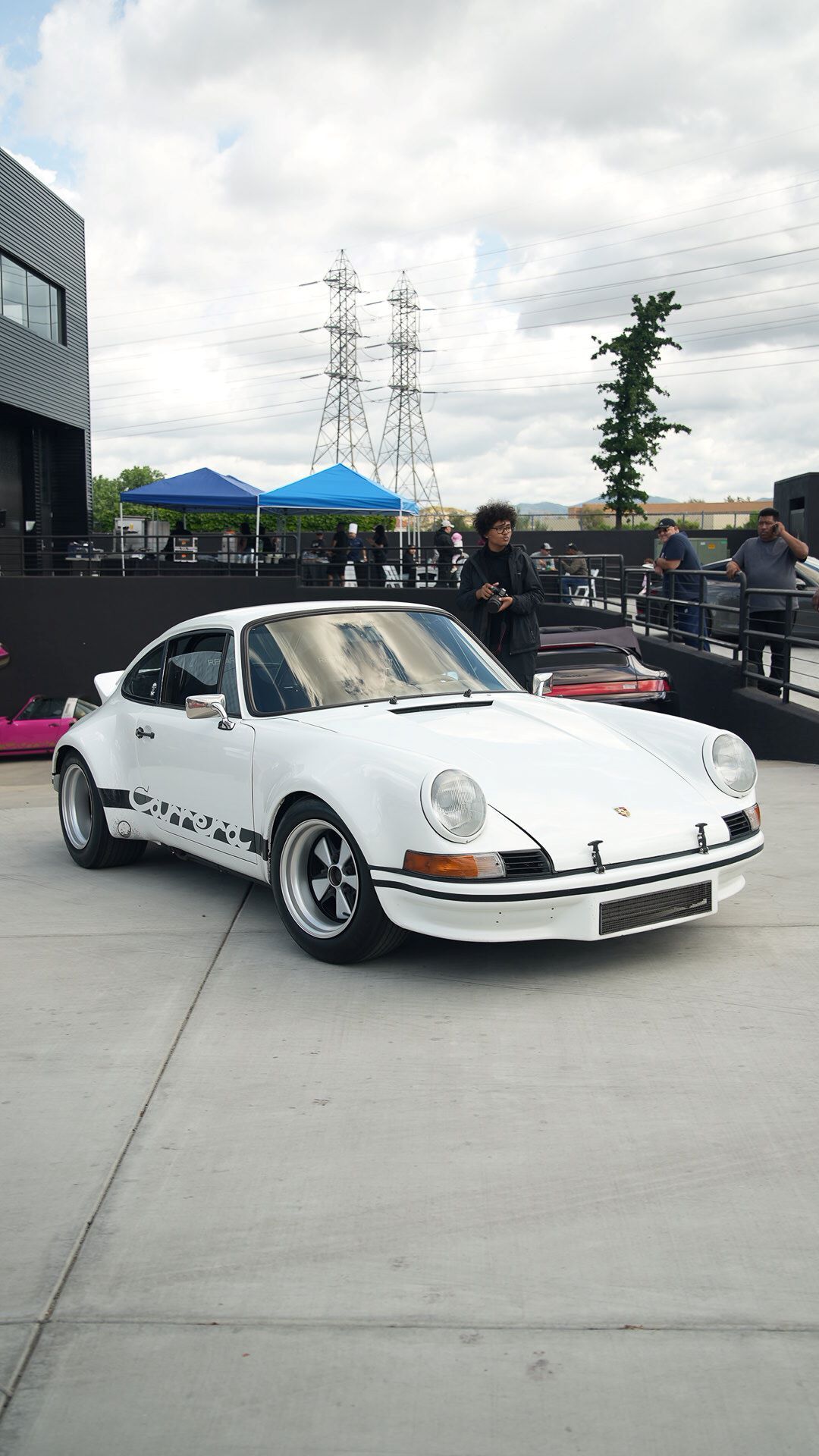 White vintage Porsche 911 Carrera with a black stripe, parked on concrete with people gathered in the background.