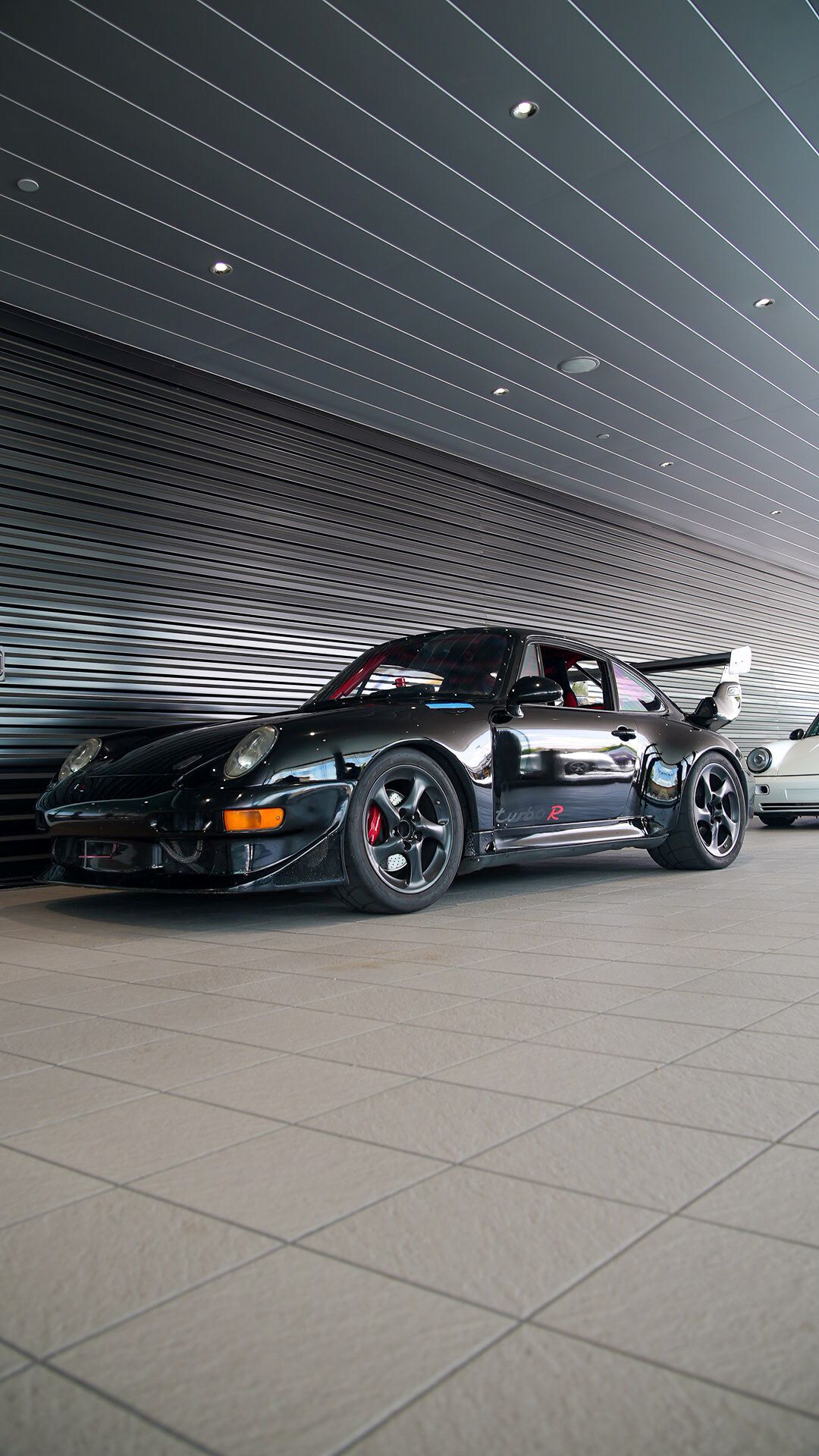 A black Porsche 911 Turbo R parked on light grey tiles beside a dark grey slatted wall.