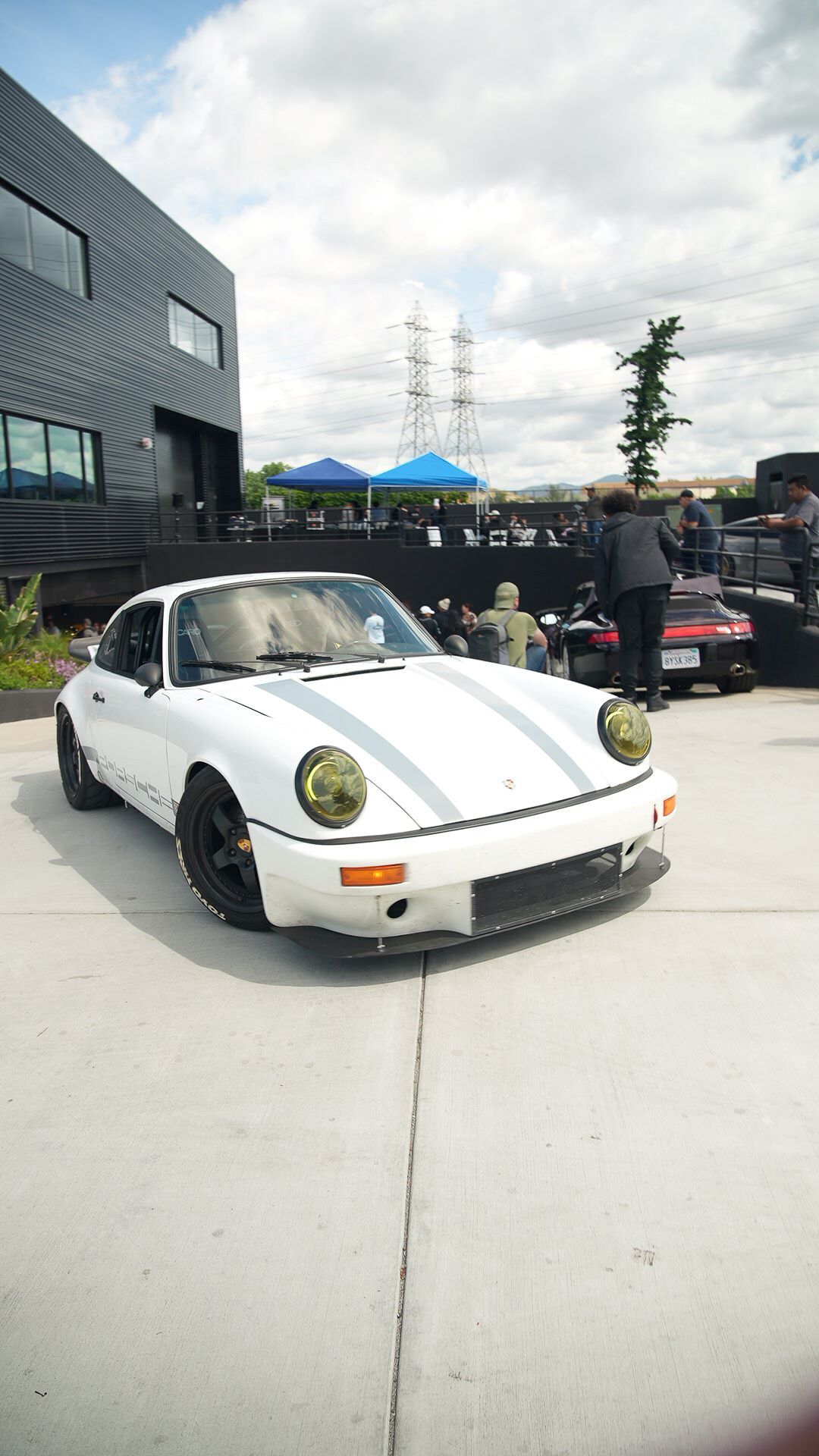 Front view of a white vintage Porsche with two gray racing stripes and yellow tinted headlights, parked outside.